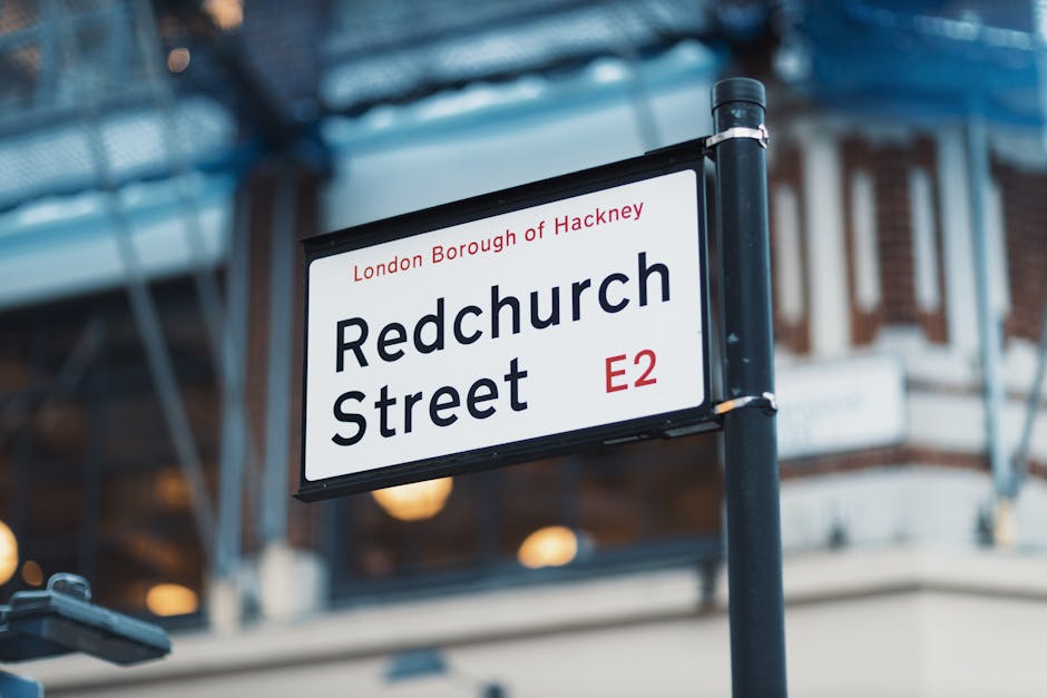 A street sign for Redchurch Street in the London Borough of Hackney, E2 postcode, mounted on a black metal pole with a blurred urban background. The sign features white background with black text displaying the street name, district, and postcode, with the district name outlined in red. Part of an old brick building with traditional architecture and a modern glass structure is visible behind the sign. The sign is illuminated by natural daylight, indicating an outdoor setting. This image is relevant to house removals and relocation services by highlighting a typical London street environment, suitable for illustrating local moving and transport processes with Man with Van Hackney providing professional removals solutions in Hackney and surrounding areas.