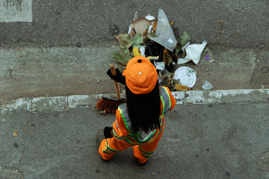 A worker dressed in high-visibility orange and green safety clothing, including a matching orange safety helmet, is standing on a grey pavement during daylight hours. The worker is using a broom to sw