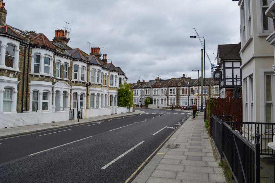 A street view in Dalston Kingsland showing a row of Victorian-style terraced houses with white façades and bay windows, situated along a paved sidewalk with black metal railings. The street has parked cars and a few pedestrians walking along the pavement. Overhead, there are streetlights and a crane visible in the background, indicating ongoing construction. The sky is overcast with grey clouds, and the scene captures a calm, residential area in Hackney. The image depicts an environment suitable for house removals and furniture transport, with clear space for loading and unloading on the street near the property entrances, which are accessible from the sidewalk. Occasionally, Man with Van Hackney provides moving and packing services in this area, supporting home relocation tasks efficiently.