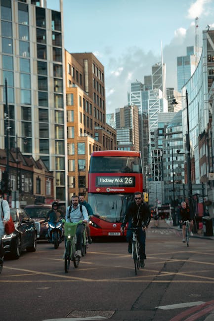 The image shows a busy urban street scene in Hackney during daytime, with tall modern office buildings and residential apartments lining the background. In the foreground, a red double-decker bus labeled 'Hackney Wick 26' is approaching, surrounded by pedestrians and cyclists crossing the street. Some individuals are walking while others are riding bikes, with a few wearing backpacks. The street is filled with parked cars on one side, and traffic lights and street signs are visible. The scene reflects typical city transport and movement, relevant to house relocations and moving logistics in the Hackney area, linking to services provided by Man with Van Hackney for removals and furniture transport in Dalston Kingsland.