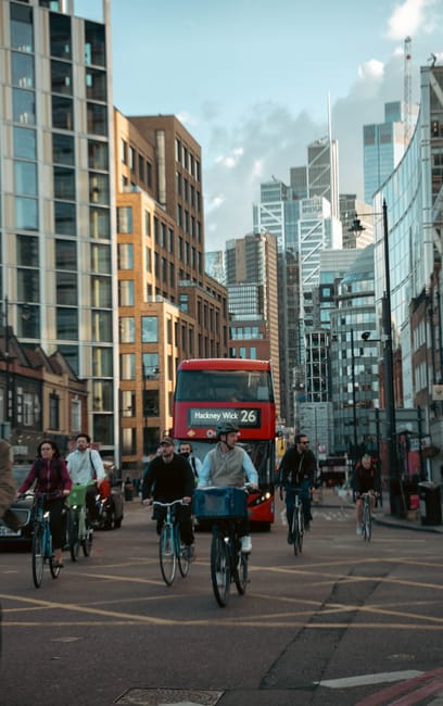 A busy urban street scene in central London with several pedestrians and cyclists moving along the roadway, including a group of five cyclists in casual clothing riding their bikes towards the camera, with some carrying bags or backpacks. Behind them, a red double-decker bus displays the route number 26 heading towards Hackney Wick, situated among tall modern office buildings made of glass and steel, as well as older brick structures. The street is busy with cars and other cyclists, and the environment is well-lit with natural daylight and partly cloudy skies overhead. This scene illustrates typical city moving logistics involving street-level transport and pedestrian activity, relevant to house removals and furniture transport services provided by companies like Man with Van Hackney, which specialise in home relocation and packing and moving processes within busy city environments.