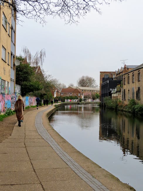 A daytime view of a paved riverside walkway alongside a narrow waterway in Hackney, with several pedestrians walking along the path. To the left, there is a building with a yellow facade and a section covered in colourful graffiti. On the right side, there are residential buildings made of brick, some with balconies and external staircases. The water reflects the overcast sky and nearby trees, some of which have sparse branches. In the background, more houses and trees are visible, creating a typical urban neighbourhood scene. The environment suggests a calm setting suitable for walking during a home relocation process, possibly involving the transportation of furniture and boxes along the river route, with Man with Van Hackney providing professional removals services for such moves.