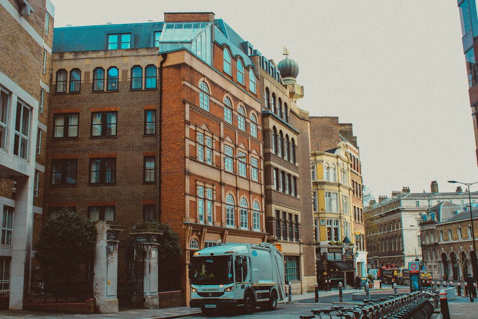 A busy urban street scene in central London with several pedestrians and cyclists moving along the roadway, including a group of five cyclists in casual clothing riding their bikes towards the camera, with some carrying bags or backpacks. Behind them, a red double-decker bus displays the route number 26 heading towards Hackney Wick, situated among tall modern office buildings made of glass and steel, as well as older brick structures. The street is busy with cars and other cyclists, and the environment is well-lit with natural daylight and partly cloudy skies overhead. This scene illustrates typical city moving logistics involving street-level transport and pedestrian activity, relevant to house removals and furniture transport services provided by companies like Man with Van Hackney, which specialise in home relocation and packing and moving processes within busy city environments.